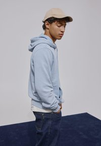 Sweat à capuche bleu clair en mélange de coton, avec poches avant et poignets côtelés, assorti à un jean bleu foncé et une casquette beige.