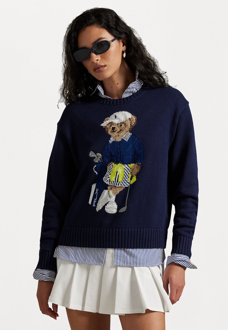 Navy knitted sweater with an embroidered bear in a golf outfit. Layered over a blue and white striped collared shirt. White pleated skirt.