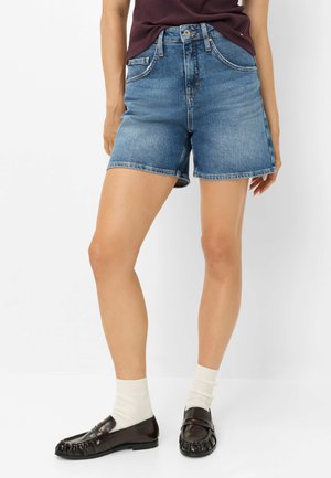Kvinde iført blå denimshorts midt på låret, mørke loafers, hvide sokker og en mørk vinrød top, der er stoppet ind, stående foran en ensfarvet hvid baggrund.