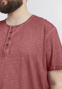 Rød, kortærmet t-shirt med rund hals og fire brune knapper. Textureret stof med et slidt udseende.