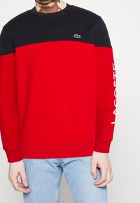 Červená a černá mikina s barevným blokovým designem, žebrovanými manžetami a bílým logem "LACOSTE" na rukávu. Bavlněný materiál.