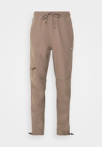 Bruine tapered joggers met elastische tailleband, trekkoord, zijzakken met rits en een klein logo nabij de linker heup. Zachte stoftextuur.