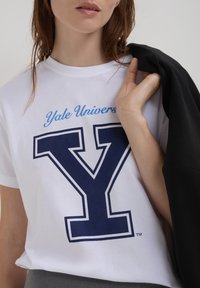 Donna con una maglietta bianca dell'Università di Yale che tiene una giacca nera sulla spalla, inquadrata dalla parte del naso verso il basso.
