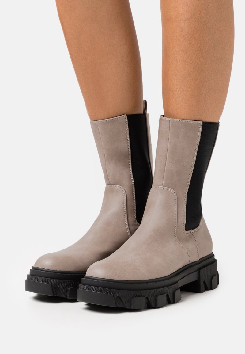 Femme portant des bottes en cuir grises mi-mollet avec des empiècements latéraux élastiques noirs et des semelles épaisses à crampons noirs, debout sur un fond blanc.