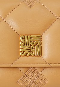 Tan lædertaske med diamantskåret quiltet tekstur, guld "SM" metallisk logo og indviklet syet mønster på overfladen.