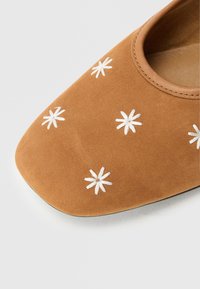 Zapato de ante color beige con puntera redonda. Presenta flores blancas bordadas dispersas en la parte superior. Textura suave y diseño minimalista.