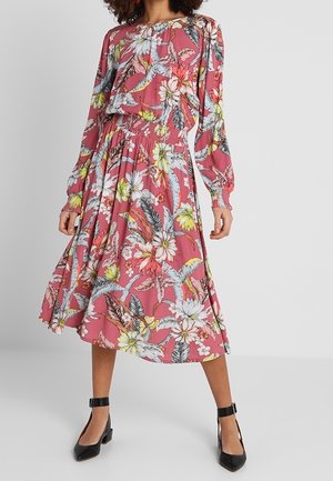 Femme portant une robe midi rose à fleurs à manches longues avec taille froncée et talons noirs à bride à la cheville, debout devant un fond uni.