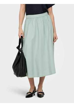 Femme portant une jupe midi vert clair, un haut noir, des escarpins slingback noirs vernis, tenant un grand sac à main noir avec un détail en forme de nœud.