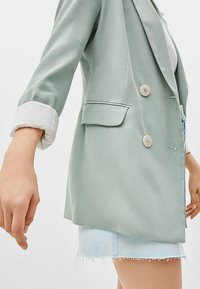 Světlý zelený blazer vyrobený z hladké látky, má dvojité zapínání, dvě boční kapsy a bílé detaily na rukávech. Nositý přes bílý top a světlé dením šortky.