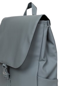 Szary plecak wykonany z gładkiego materiału syntetycznego, z klapą, regulowanym paskiem i boczną kieszenią na dodatkowe przechowywanie.