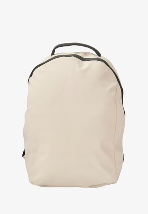 Mochila de color beige claro con una textura suave y mate, forma redondeada, acentos negros en contraste y un logo estampado en la parte delantera.