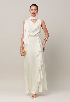 Femme portant une robe ivoire sans manches avec un détail volanté, un bracelet en or, des boucles d'oreilles, et tenant une pochette dorée texturée.