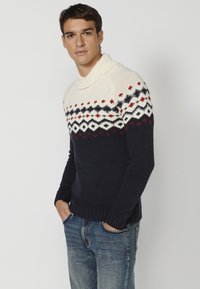 Jersey de cuello alto de punto que presenta un patrón geométrico en crema, azul marino y rojo. Puños y dobladillo acanalados, hecho de hilo texturizado para mayor calidez.