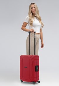 Roter Hartschalenkoffer mit texturer Oberfläche, schwarzem Teleskopgriff und Rollen. Verfügt über ein integriertes Schloss und ein schräges Design.