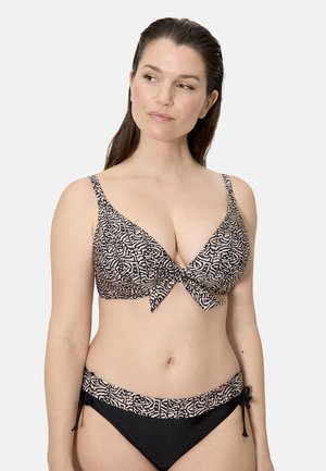 Mujer con un bikini estampado en negro y beige, con un top con lazo delantero y braguitas a juego con lazos laterales, sobre un fondo liso.