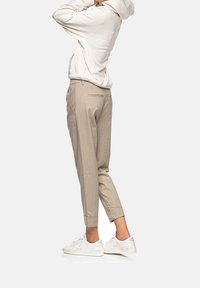 Beżowe spodnie na miarę z mankietem u dołu, o gładkiej fakturze i dwoma tylnymi kieszeniami, w zestawie z jasną bluzą z kapturem i trampkami.
