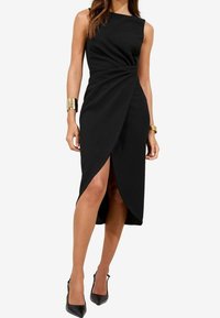 Kvinna klädd i en ärmlös svart omslagsklänning med hög slit och svarta spetsiga klackskor, smyckad med guldfärgade manschettarmband.