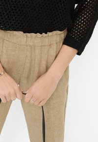 Pantaloni di lino beige con vita arricciata, dotati di tasche laterali, abbinati a un maglione nero a maglia aperta. Mani tengono un cordino nero.