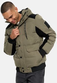 Veste rembourrée en vert olive avec des accents noirs. Dispose d'une capuche, de poches avant et de poignets côtelés. Texture douce avec des détails en design matelassé.