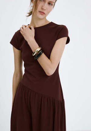 Donna che indossa un abito marrone scuro a maniche corte e bracciali spessi neri e dorati, posa con la mano vicino al collo su uno sfondo semplice.