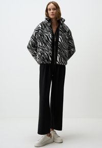 Zwart-witte nepbont jas met een zebrapatroon, hoge kraag en ritssluiting, gedragen over een zwarte outfit en witte sneakers.