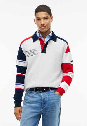 Junger Mann trägt ein farblich abgesetztes Poloshirt mit roten, weißen und marineblauen Streifen und hellblauen Jeans, steht mit einer Hand in der Tasche.