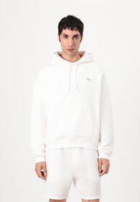 Sudadera blanca de tejido suave, con un bolsillo tipo canguro y un pequeño logo en el pecho, combinada con pantalones cortos blancos texturizados.