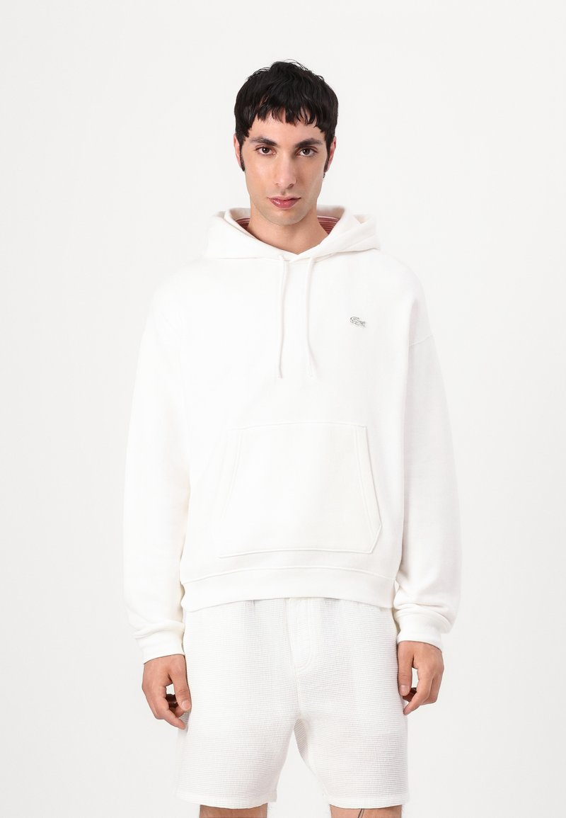 Sudadera blanca de tejido suave, con un bolsillo tipo canguro y un pequeño logo en el pecho, combinada con pantalones cortos blancos texturizados.
