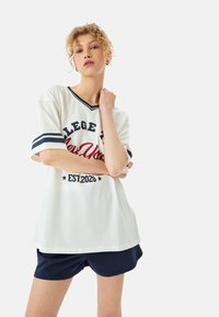 Camiseta blanca de gran tamaño con acentos en navy y texto "COLLEGE NY EST. 2020" combinada con pantalones cortos navy; diseño de mangas cortas y cuello en V.