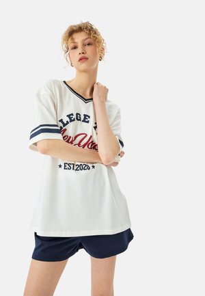 T-shirt bianco oversize con accenti blu navy e testo "COLLEGE NY EST. 2020" indossato con pantaloni corti blu navy; maniche corte e design a V.