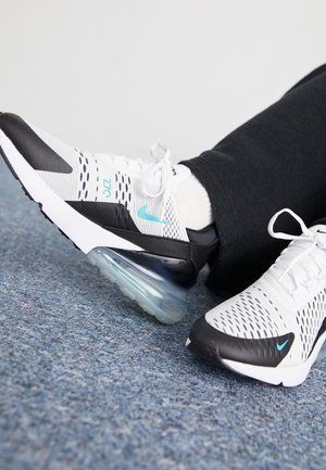 Personne portant des baskets Nike Air Max 270 blanches et noires avec le logo Nike swoosh bleu clair, assise sur un sol recouvert de moquette bleue.
