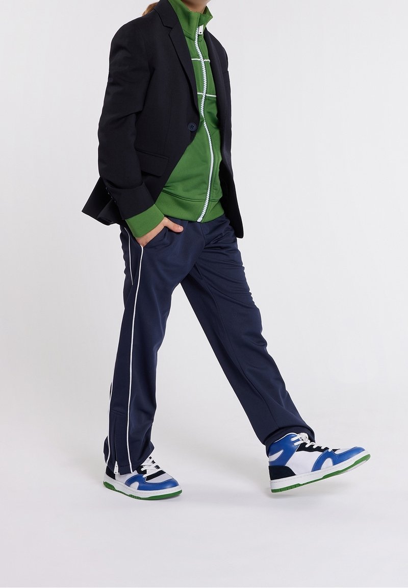 Personne marchant, portant un blazer noir, une veste zippée verte, un pantalon de survêtement bleu marine avec des rayures blanches, et des baskets bleues et blanches avec des semelles vertes.