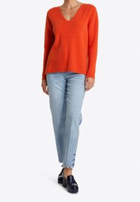 Pull orange côtelé avec un col en V, associé à un jean droit bleu clair. Le mannequin porte des mocassins noirs épais.