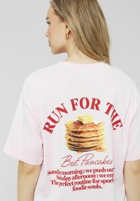 T-shirt surdimensionné rose en coton, arborant un graphique de pancakes avec du sirop et du beurre, du texte rouge et des manches courtes retroussées.