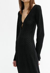 Schwarzes langärmliges Kleid mit tiefem V-Ausschnitt und Schlüssellochdetail, das ein strukturiertes Muster im gesamten Stoff aufweist.