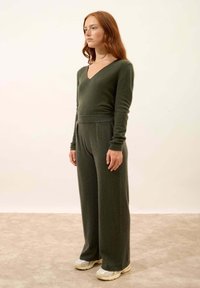 Ciemnozielony, dopasowany sweter z dekoltem w serek, w zestawieniu z szerokimi spodniami w dopasowanym kolorze. Model nosi beżowe sneakersy. Gładka tekstura.