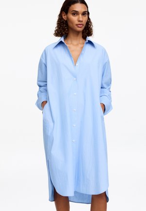 Femme portant une robe chemisier ample bleu clair à manches longues, avec col et poches, debout les mains dans les poches.