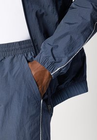 Marine vindjakke med elastiske manchetter og hvide kantsting. Har en lynlåslomme og tekstureret stof, parret med en tætsiddende hvid skjorte.