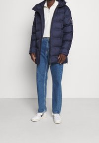 Giubbotto imbottito blu navy con zip completa, cappuccio e polsini a costine, abbinato a jeans a gamba diritta azzurri chiari e sneakers bianche.