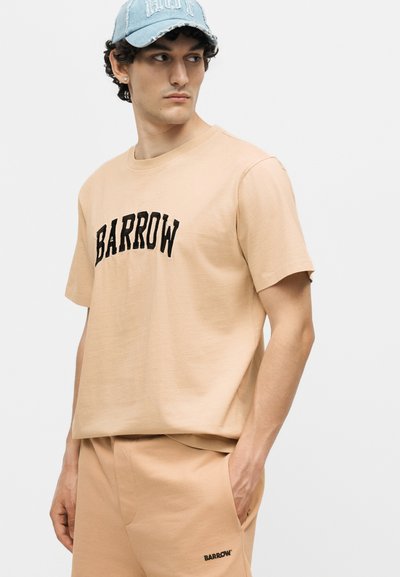 Νέος άνδρας που φοράει μπεζ μπλουζάκι Barrow και ταιριαστό σορτς, με ανοιχτό μπλε τζιν καπέλο, κοιτάζει προς τα αριστερά.