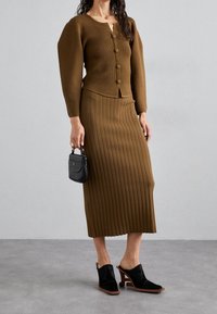 Kvinna iklädd brun ribbad stickad kjol och matchande knappad kofta, håller en liten svart handväska och har på sig svarta klackiga mules.