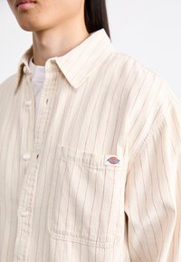 Beige Hemd mit roten vertikalen Streifen, ausgestattet mit einer Brusttasche und einem kleinen Logo-Patch. Hergestellt aus Baumwolle, mit klassischem Kragen und Knopfmanschetten.