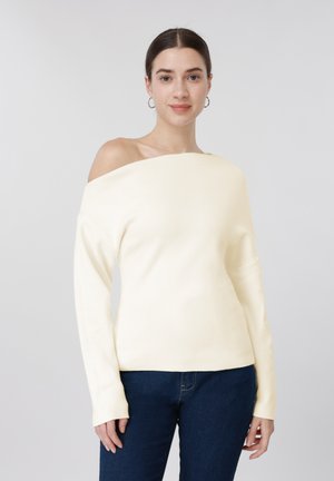 Žena v krémovém svetru s odhalenými rameny a tmavě modrých džínách stojí před jednoduchým světle šedým pozadím.