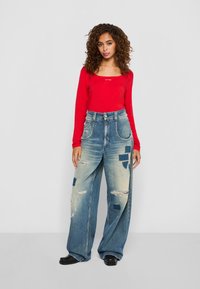 Vrouw met krullend haar, gekleed in een rode lange mouwen top, losse wijde jeans met patches en scheuren, en zwarte schoenen, staande tegen een eenvoudige achtergrond.