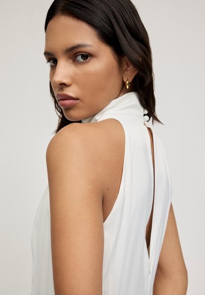 Vestido blanco sin mangas con cuello alto y espalda descubierta. Presenta una textura suave y botones decorativos en el cuello. Diseño minimalista.