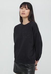 Felpa nera con testo in rilievo "AMOUR", colletto a giro, spalle drop, e polsini a coste. Il tessuto sembra morbido con una finitura opaca.