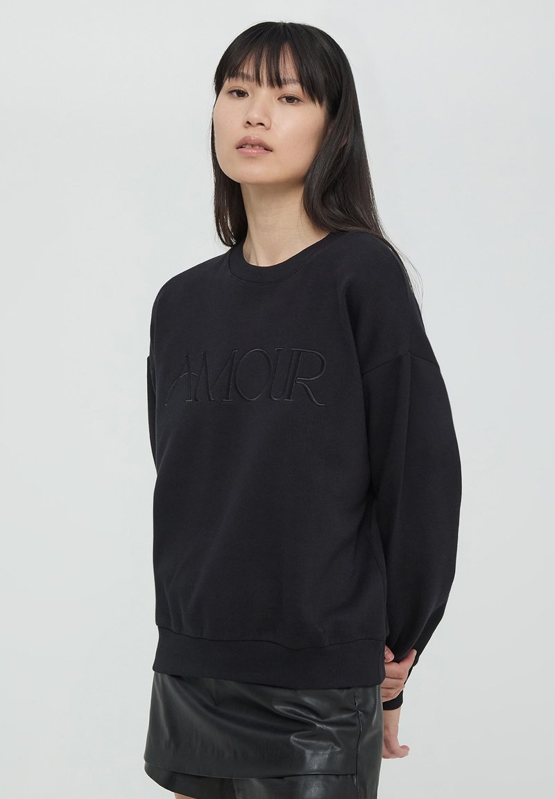 Felpa nera con testo in rilievo "AMOUR", colletto a giro, spalle drop, e polsini a coste. Il tessuto sembra morbido con una finitura opaca.