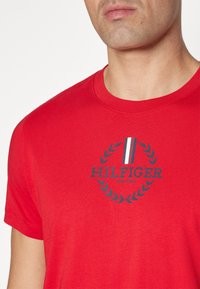 Rød bomulds t-shirt med rund halsudskæring, korte ærmer og et marineblåt cirkulært logo med en laurbærkrans og stribede detaljer.