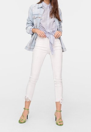 Vrouw draagt een lichtblauw spijkerjasje, een geknoopte lichtblauwe blouse, witte cropped jeans met rafelige zomen en groene sandalen met hoge hakken, staand.