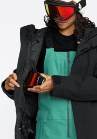 Schwarze isolierte Jacke über einem schwarzen Shirt, grüne Skihose und rot getönte Skibrille in der Hand. Hat Reißverschlüsse und Klettverschluss-Details.